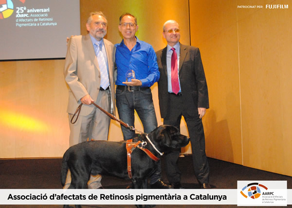 Peu de foto, logo de l’Associació d’Afectats de Retinosis Pigmentària a Catalunya. Fotografia del 25è aniversari de l'associació fent entrega del premi al Dr. Josep Torrent al mig del Sr. Jordi Palà amb el seu gos pigall i el Sr. Albert Espanyol.